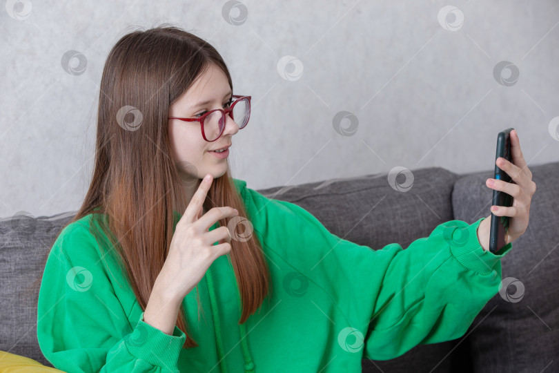 Скачать Подросток в зеленой толстовке с капюшоном делает селфи, сидя днем на сером диване в уютном помещении фотосток Ozero