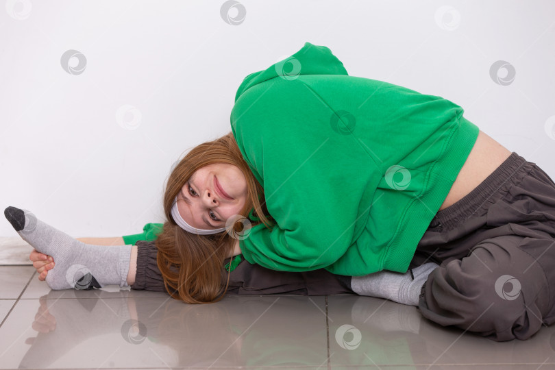 Скачать Девушка-подросток в зеленой толстовке демонстрирует впечатляющую гибкость, потягиваясь в светлой комнате во время занятия йогой фотосток Ozero