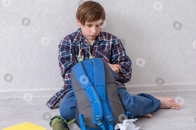 Скачать Маленький мальчик-школьник сидит дома на полу и распаковывает свой рюкзак. Ему грустно и скучно. фотосток Ozero