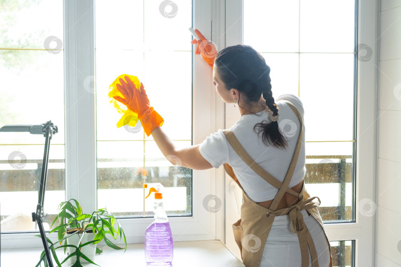 Скачать Женщина в резиновых перчатках и фартуке вручную моет окно балкона дома с помощью тряпки для мытья посуды и швабры внутри помещения. Наведение весеннего порядка и чистоты, услуги по уборке фотосток Ozero
