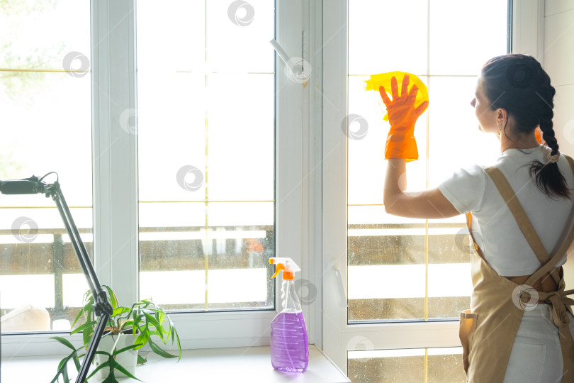 Скачать Женщина в резиновых перчатках и фартуке вручную моет окно балкона дома с помощью тряпки для мытья посуды и швабры внутри помещения. Наведение весеннего порядка и чистоты, услуги по уборке фотосток Ozero