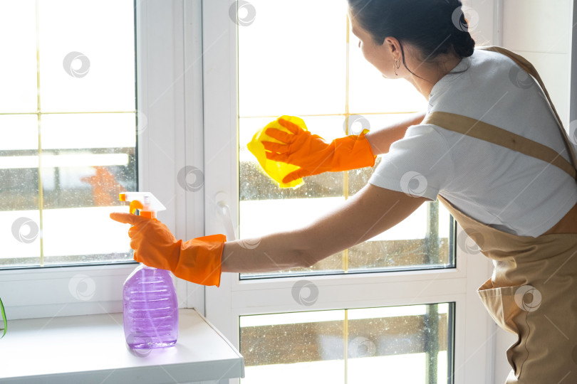 Скачать Женщина в резиновых перчатках и фартуке вручную моет окно балкона дома с помощью тряпки для мытья посуды и швабры внутри помещения. Наведение весеннего порядка и чистоты, услуги по уборке фотосток Ozero