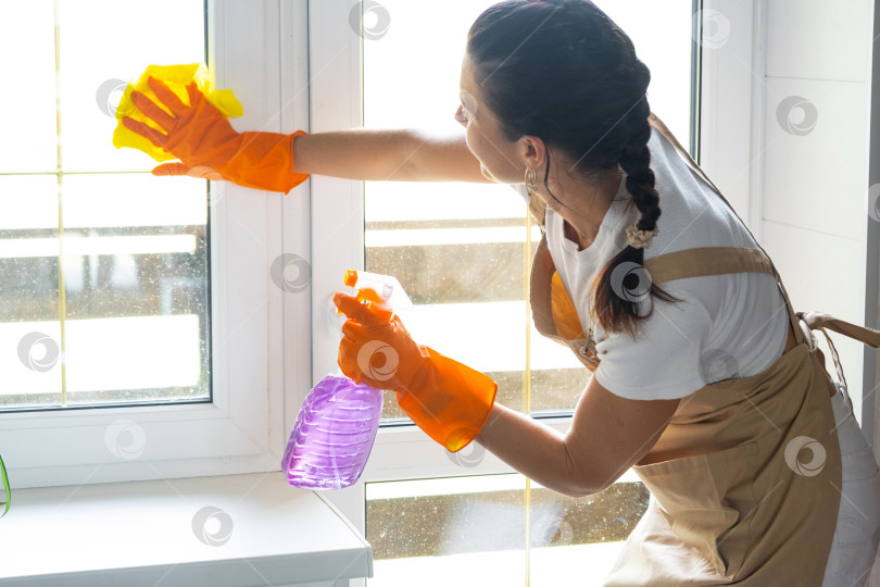 Скачать Женщина в резиновых перчатках и фартуке вручную моет окно балкона дома с помощью тряпки для мытья посуды и швабры внутри помещения. Наведение весеннего порядка и чистоты, услуги по уборке фотосток Ozero