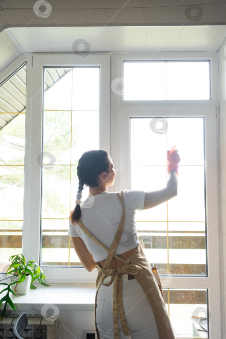 Скачать Женщина в резиновых перчатках и фартуке вручную моет окно балкона дома с помощью тряпки для мытья посуды и швабры внутри помещения. Наведение весеннего порядка и чистоты, услуги по уборке фотосток Ozero