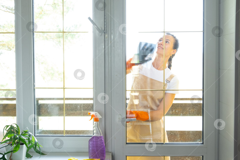 Скачать Женщина в резиновых перчатках и фартуке вручную моет балконное окно дома тряпкой для мытья посуды и шваброй снаружи, за стеклом. Наведение весеннего порядка и чистоты, услуги по уборке фотосток Ozero