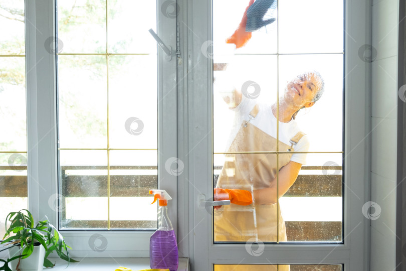 Скачать Женщина в резиновых перчатках и фартуке вручную моет балконное окно дома тряпкой для мытья посуды и шваброй снаружи, за стеклом. Наведение весеннего порядка и чистоты, услуги по уборке фотосток Ozero