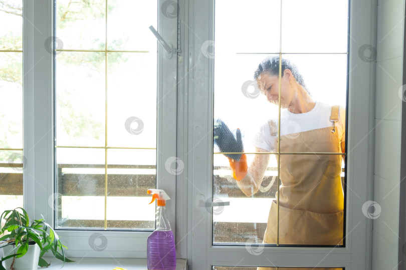Скачать Женщина в резиновых перчатках и фартуке вручную моет балконное окно дома тряпкой для мытья посуды и шваброй снаружи, за стеклом. Наведение весеннего порядка и чистоты, услуги по уборке фотосток Ozero