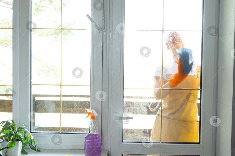 Скачать Женщина в резиновых перчатках и фартуке вручную моет балконное окно дома тряпкой для мытья посуды и шваброй снаружи, за стеклом. Наведение весеннего порядка и чистоты, услуги по уборке фотосток Ozero