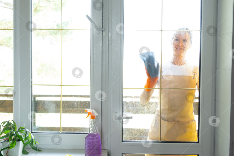 Скачать Женщина в резиновых перчатках и фартуке вручную моет балконное окно дома тряпкой для мытья посуды и шваброй снаружи, за стеклом. Наведение весеннего порядка и чистоты, услуги по уборке фотосток Ozero