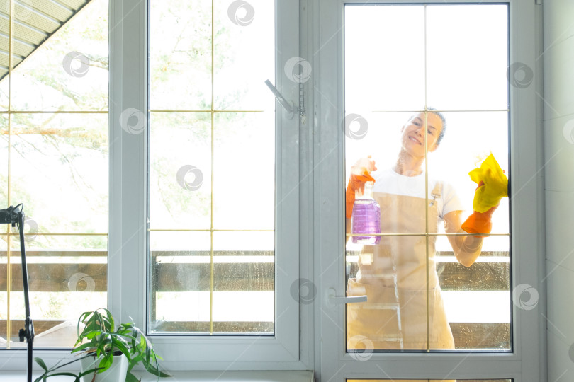 Скачать Женщина в резиновых перчатках и фартуке вручную моет балконное окно дома тряпкой для мытья посуды и шваброй снаружи, за стеклом. Наведение весеннего порядка и чистоты, услуги по уборке фотосток Ozero
