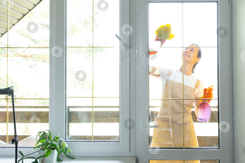 Скачать Женщина в резиновых перчатках и фартуке вручную моет балконное окно дома тряпкой для мытья посуды и шваброй снаружи, за стеклом. Наведение весеннего порядка и чистоты, услуги по уборке фотосток Ozero