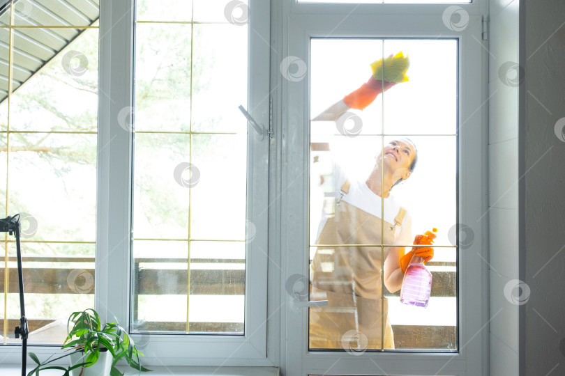 Скачать Женщина в резиновых перчатках и фартуке вручную моет балконное окно дома тряпкой для мытья посуды и шваброй снаружи, за стеклом. Наведение весеннего порядка и чистоты, услуги по уборке фотосток Ozero
