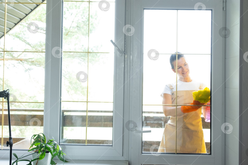 Скачать Женщина в резиновых перчатках и фартуке вручную моет балконное окно дома тряпкой для мытья посуды и шваброй снаружи, за стеклом. Наведение весеннего порядка и чистоты, услуги по уборке фотосток Ozero