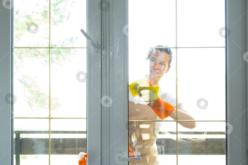 Скачать Женщина в резиновых перчатках и фартуке вручную моет балконное окно дома тряпкой для мытья посуды и шваброй снаружи, за стеклом. Наведение весеннего порядка и чистоты, услуги по уборке фотосток Ozero