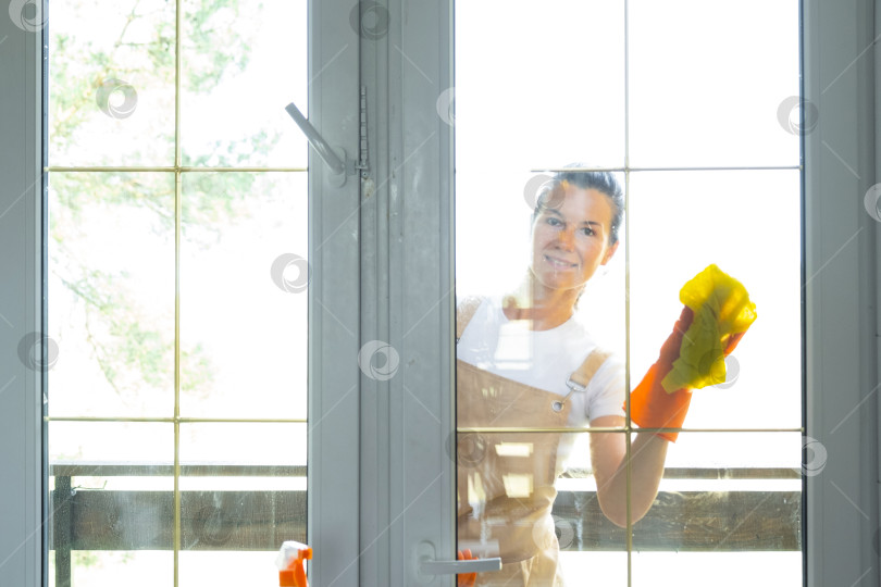 Скачать Женщина в резиновых перчатках и фартуке вручную моет балконное окно дома тряпкой для мытья посуды и шваброй снаружи, за стеклом. Наведение весеннего порядка и чистоты, услуги по уборке фотосток Ozero