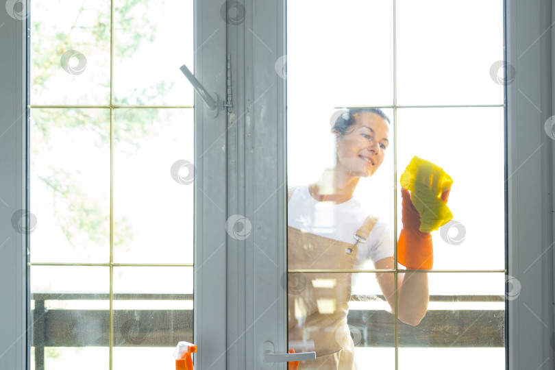 Скачать Женщина в резиновых перчатках и фартуке вручную моет балконное окно дома тряпкой для мытья посуды и шваброй снаружи, за стеклом. Наведение весеннего порядка и чистоты, услуги по уборке фотосток Ozero