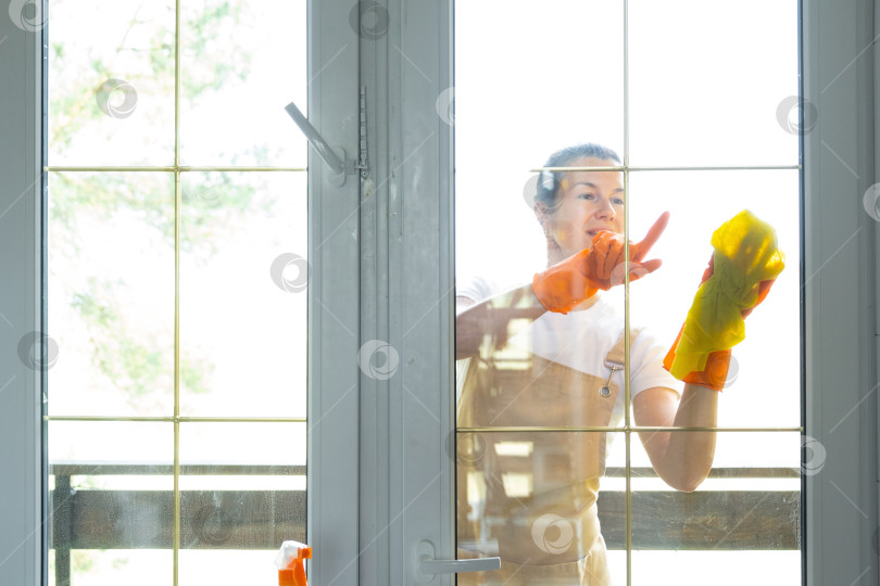 Скачать Женщина в резиновых перчатках и фартуке вручную моет балконное окно дома тряпкой для мытья посуды и шваброй снаружи, за стеклом. Наведение весеннего порядка и чистоты, услуги по уборке фотосток Ozero