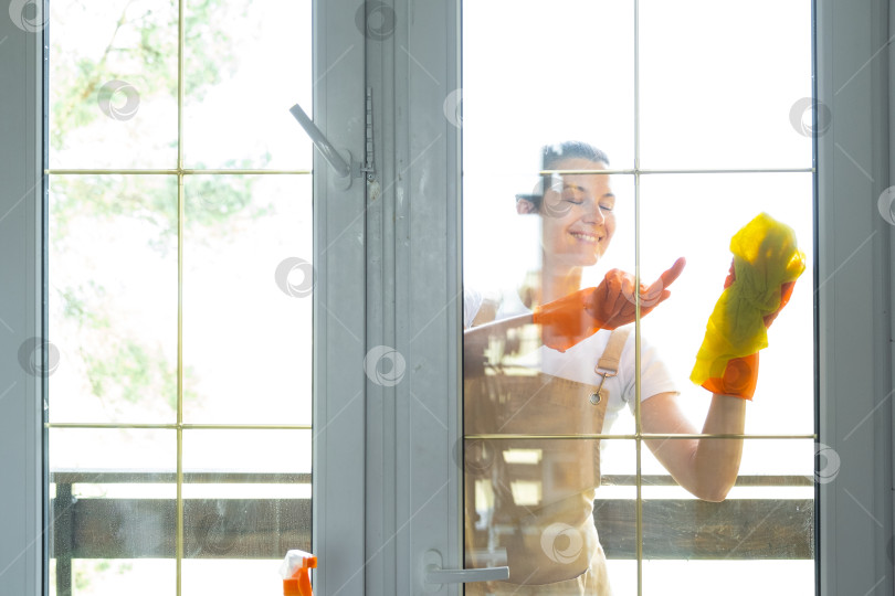 Скачать Женщина в резиновых перчатках и фартуке вручную моет балконное окно дома тряпкой для мытья посуды и шваброй снаружи, за стеклом. Наведение весеннего порядка и чистоты, услуги по уборке фотосток Ozero