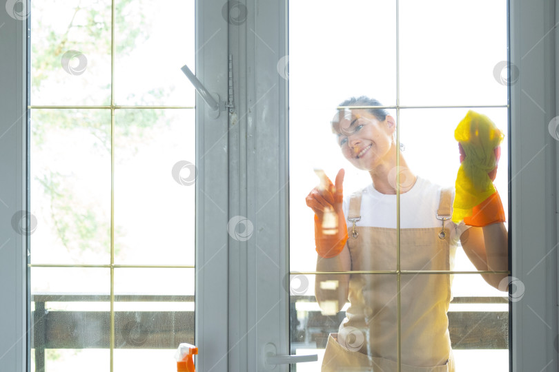 Скачать Женщина в резиновых перчатках и фартуке вручную моет балконное окно дома тряпкой для мытья посуды и шваброй снаружи, за стеклом. Наведение весеннего порядка и чистоты, услуги по уборке фотосток Ozero