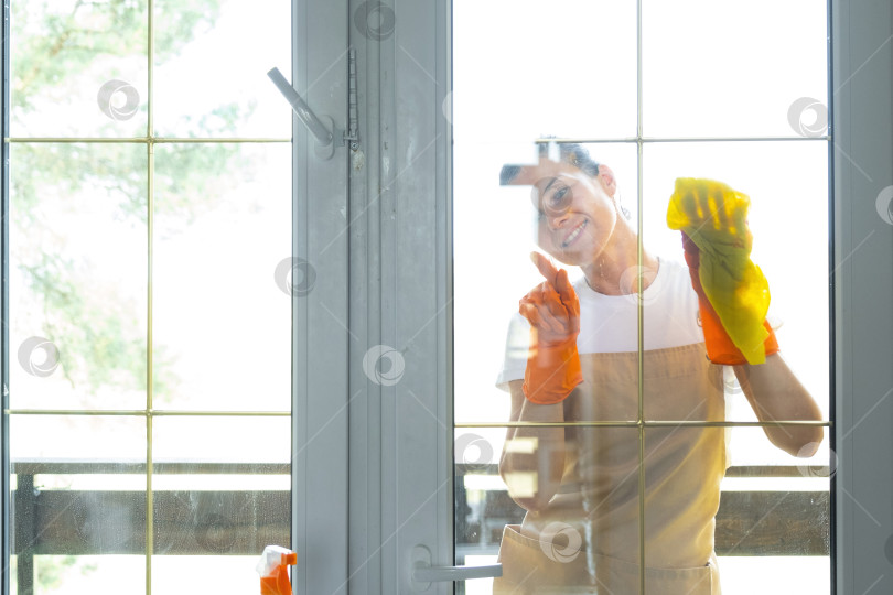 Скачать Женщина в резиновых перчатках и фартуке вручную моет балконное окно дома тряпкой для мытья посуды и шваброй снаружи, за стеклом. Наведение весеннего порядка и чистоты, услуги по уборке фотосток Ozero