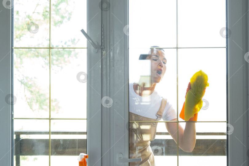 Скачать Женщина в резиновых перчатках и фартуке вручную моет балконное окно дома тряпкой для мытья посуды и шваброй снаружи, за стеклом. Наведение весеннего порядка и чистоты, услуги по уборке фотосток Ozero