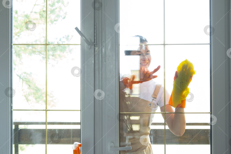 Скачать Женщина в резиновых перчатках и фартуке вручную моет балконное окно дома тряпкой для мытья посуды и шваброй снаружи, за стеклом. Наведение весеннего порядка и чистоты, услуги по уборке фотосток Ozero