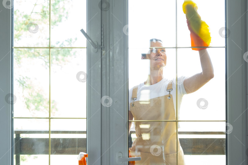 Скачать Женщина в резиновых перчатках и фартуке вручную моет балконное окно дома тряпкой для мытья посуды и шваброй снаружи, за стеклом. Наведение весеннего порядка и чистоты, услуги по уборке фотосток Ozero