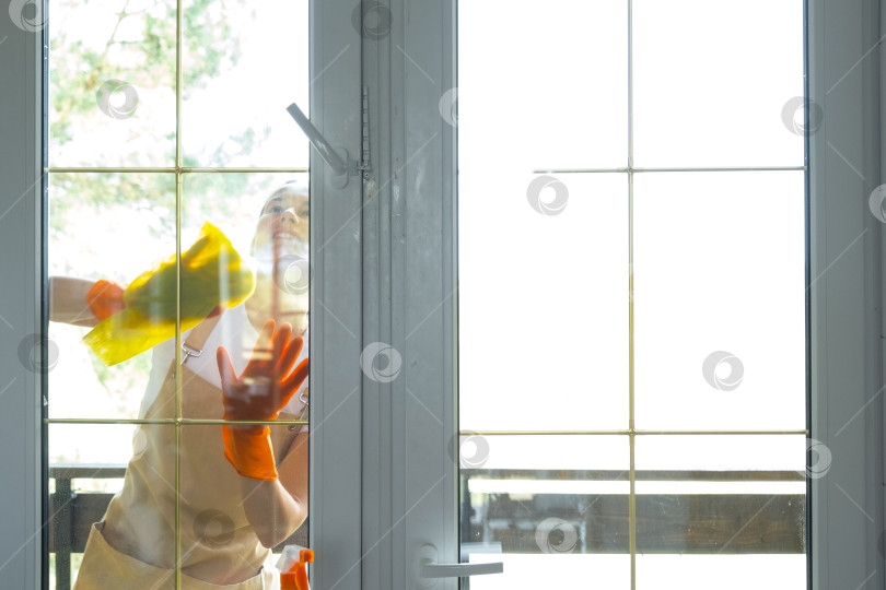 Скачать Женщина в резиновых перчатках и фартуке вручную моет балконное окно дома тряпкой для мытья посуды и шваброй снаружи, за стеклом. Наведение весеннего порядка и чистоты, услуги по уборке фотосток Ozero