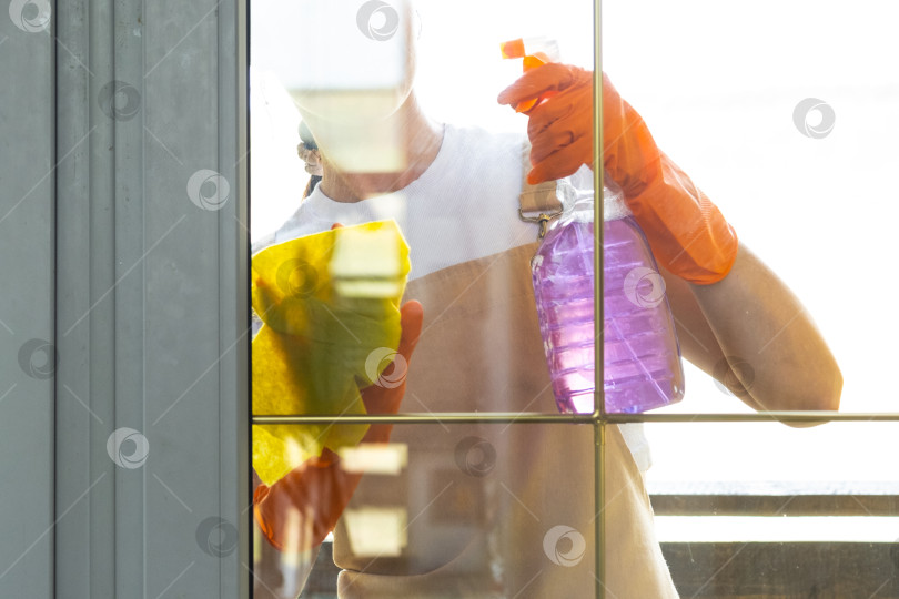Скачать Женщина в резиновых перчатках и фартуке вручную моет балконное окно дома тряпкой для мытья посуды и шваброй снаружи, за стеклом. Наведение весеннего порядка и чистоты, услуги по уборке фотосток Ozero