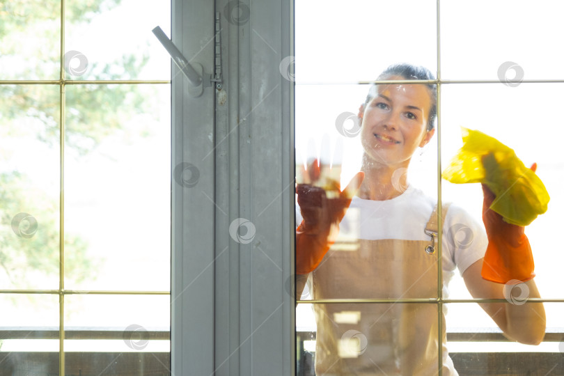 Скачать Женщина в резиновых перчатках и фартуке вручную моет балконное окно дома тряпкой для мытья посуды и шваброй снаружи, за стеклом. Наведение весеннего порядка и чистоты, услуги по уборке фотосток Ozero