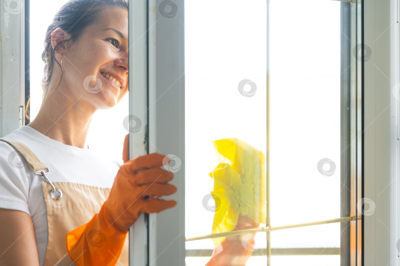 Скачать Женщина в резиновых перчатках и фартуке вручную моет балконное окно дома тряпкой для мытья посуды и шваброй снаружи, за стеклом. Наведение весеннего порядка и чистоты, услуги по уборке фотосток Ozero
