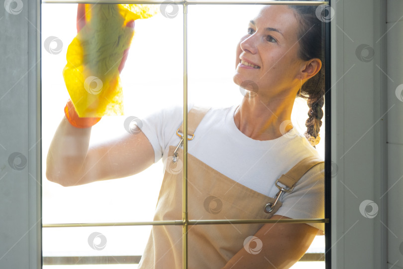 Скачать Женщина в резиновых перчатках и фартуке вручную моет балконное окно дома тряпкой для мытья посуды и шваброй снаружи, за стеклом. Наведение весеннего порядка и чистоты, услуги по уборке фотосток Ozero