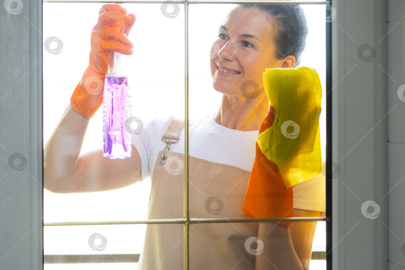 Скачать Женщина в резиновых перчатках и фартуке вручную моет балконное окно дома тряпкой для мытья посуды и шваброй снаружи, за стеклом. Наведение весеннего порядка и чистоты, услуги по уборке фотосток Ozero