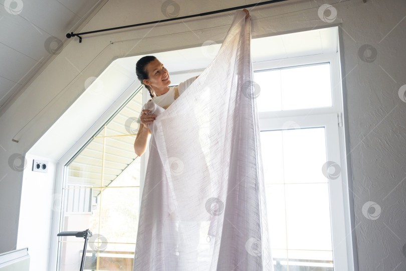 Скачать Женщина в фартуке вешает прозрачные тюлевые занавески на большие мансардные окна в доме с белым интерьером. Генеральная уборка, наведение порядка фотосток Ozero