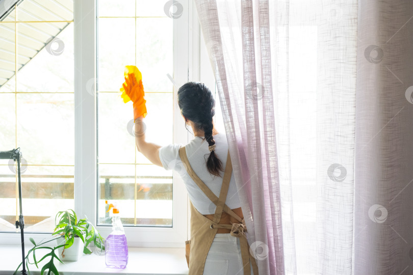 Скачать Женщина в резиновых перчатках и фартуке вручную моет окно балкона дома с помощью тряпки для мытья посуды и швабры внутри помещения. Наведение весеннего порядка и чистоты, услуги по уборке фотосток Ozero