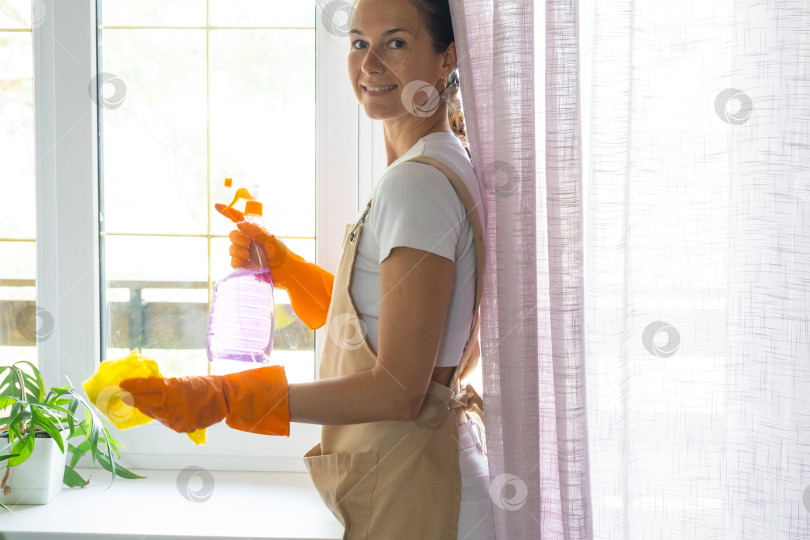 Скачать Женщина в резиновых перчатках и фартуке вручную моет окно балкона дома с помощью тряпки для мытья посуды и швабры внутри помещения. Наведение весеннего порядка и чистоты, услуги по уборке фотосток Ozero