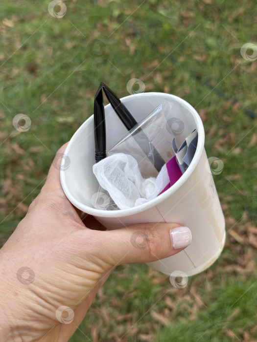 Скачать Рука держит бумажный стаканчик, наполненный разнообразным пластиковым мусором, на фоне травы фотосток Ozero