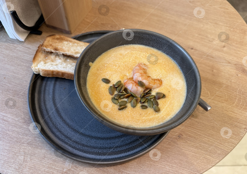 Скачать Тыквенный суп с креветками и тыквенными семечками на тарелке с тостами, подается на деревянном столе фотосток Ozero