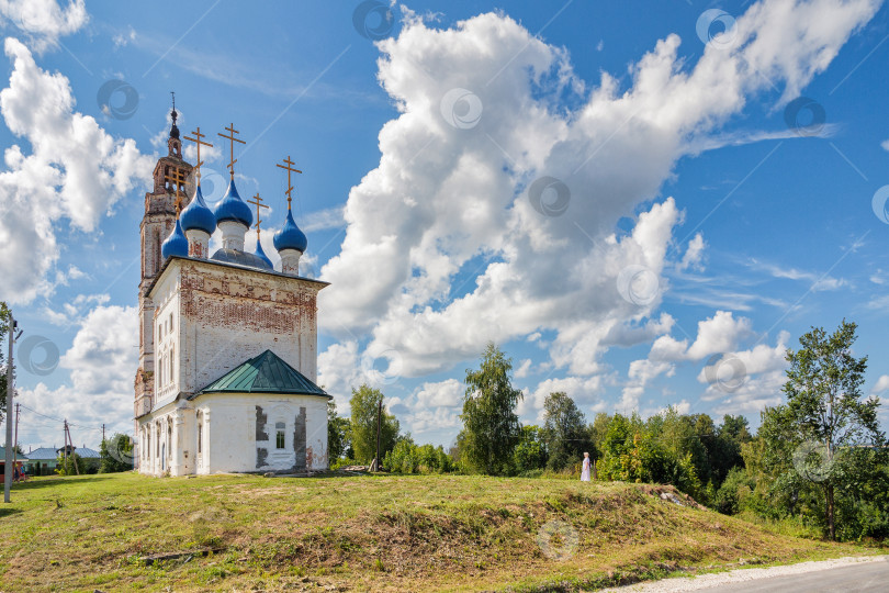 Скачать Старинная белокаменная Покровская церковь с колокольней в селе Клязьминский Городок (Стародуб). На фоне видны голубое небо и большие белые облака. На лужайке виден силуэт женщины. фотосток Ozero