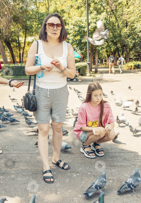 Скачать Молодая испаноязычная девушка сидит на корточках возле стаи голубей в городском парке. Рядом стоит взрослая испаноязычная женщина, и они оба наслаждаются оживленной атмосферой. фотосток Ozero