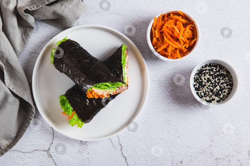 Скачать Тунец, сыр, корейская морковь и листья салата, завернутые в нори, на тарелке на столе, вид сверху фотосток Ozero