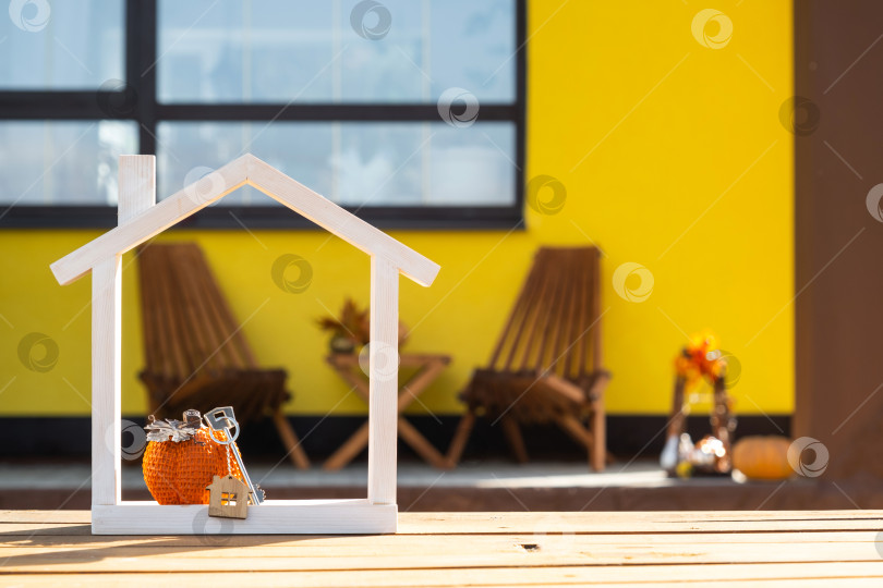 Скачать Осенний декор на крыльце желтого дома в виде тыквы, фонаря, гирлянд и фигурки крошечного домика и ключа. Вечеринка в честь Хэллоуина, осеннее настроение, праздник урожая, недвижимость, страхование, ипотека фотосток Ozero