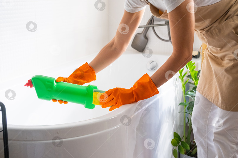 Скачать Женщина в резиновых перчатках и фартуке вручную моет ванну в ванной комнате дома губкой для мытья посуды и чистящим средством во флаконе. Наведение порядка, чистоты и гигиены в доме, услуги по уборке фотосток Ozero