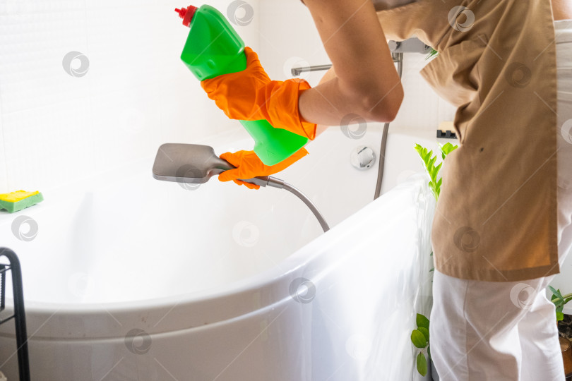 Скачать Женщина в резиновых перчатках и фартуке вручную моет ванну в ванной комнате дома с помощью губки для мытья посуды и чистящего средства в бутылочке. Наведение порядка, чистоты и гигиены в доме, услуги по уборке фотосток Ozero
