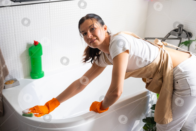 Скачать Женщина в резиновых перчатках и фартуке вручную моет ванну в ванной комнате дома губкой для мытья посуды и чистящим средством во флаконе. Наведение порядка, чистоты и гигиены в доме, услуги по уборке фотосток Ozero