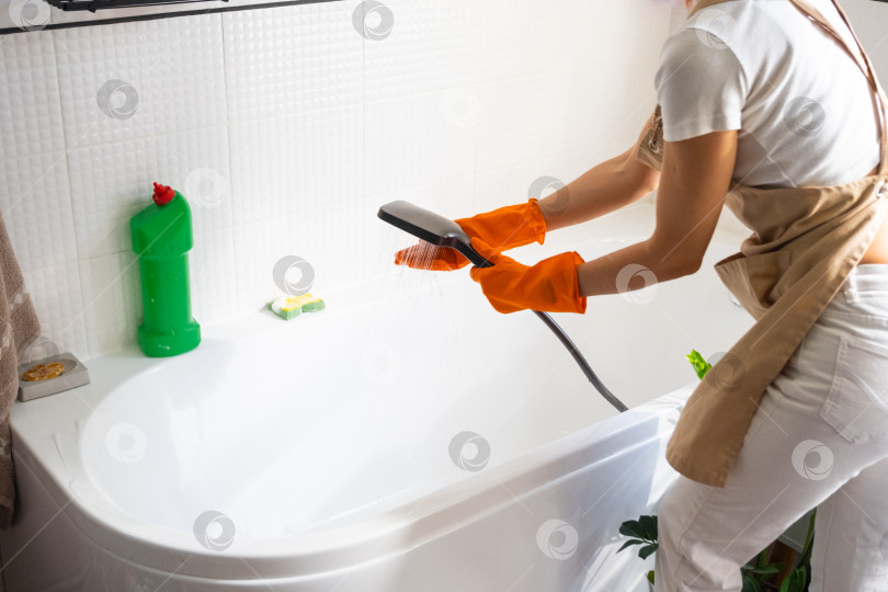 Скачать Женщина в резиновых перчатках и фартуке вручную моет ванну в ванной комнате дома с помощью губки для мытья посуды и чистящего средства в бутылочке. Наведение порядка, чистоты и гигиены в доме, услуги по уборке фотосток Ozero