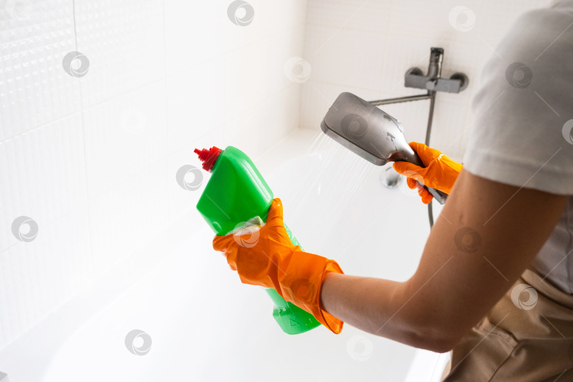 Скачать Женщина в резиновых перчатках и фартуке вручную моет ванну в ванной комнате дома с помощью губки для мытья посуды и чистящего средства в бутылочке. Наведение порядка, чистоты и гигиены в доме, услуги по уборке фотосток Ozero