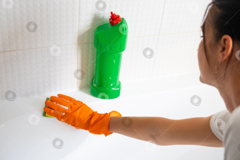Скачать Руки в резиновых перчатках крупным планом и бутылка с моющим средством в ванной комнате дома, губка для мытья посуды и чистящее средство на ванне. Наведение порядка, чистоты и гигиены в доме, услуги по уборке фотосток Ozero