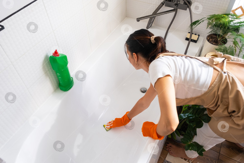 Скачать Женщина в резиновых перчатках и фартуке вручную моет ванну в ванной комнате дома с помощью губки для мытья посуды и чистящего средства в бутылочке. Наведение порядка, чистоты и гигиены в доме, услуги по уборке фотосток Ozero
