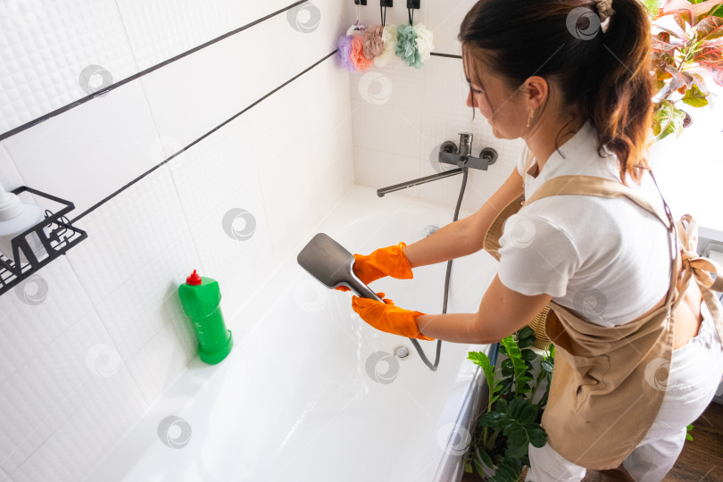 Скачать Женщина в резиновых перчатках и фартуке вручную моет ванну в ванной комнате дома губкой для мытья посуды и чистящим средством во флаконе. Наведение порядка, чистоты и гигиены в доме, услуги по уборке фотосток Ozero